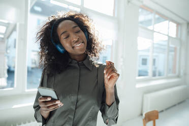 Young woman holding mobile phone enjoying music listening through wireless headphones in office - KNSF09476