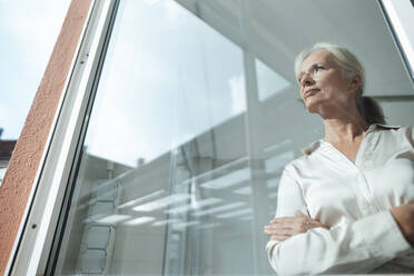 Thoughtful businesswoman standing with arms crossed seen through glass - KNSF09531