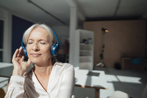 Lächelnde ältere Frau mit geschlossenen Augen hört Musik über drahtlose Kopfhörer im Büro - KNSF09538