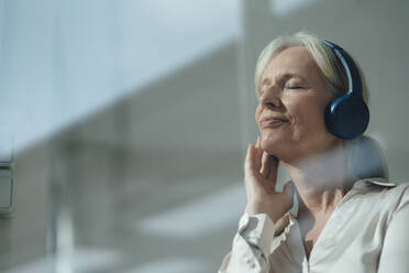 Lächelnde ältere Frau mit geschlossenen Augen hört Musik über drahtlose Kopfhörer - KNSF09540