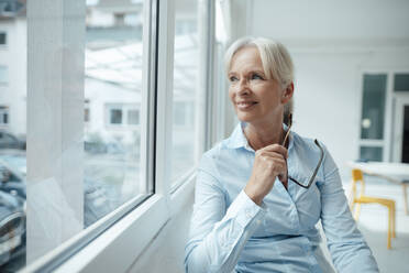 Lächelnde ältere Geschäftsfrau mit Brille schaut durch ein Fenster im Büro - KNSF09544