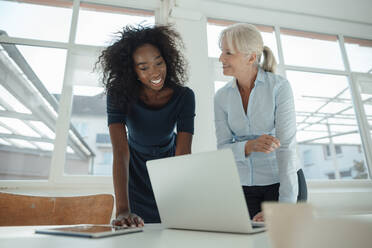 Lächelnde Geschäftsfrauen, die über einem Laptop am Schreibtisch im Büro diskutieren - KNSF09557