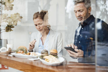 Geschäftsfrau beim Mittagessen sitzend neben einem Geschäftsmann, der ein Smartphone in einem Café benutzt, gesehen durch Glas - JOSEF10812