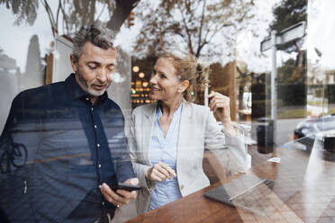 Geschäftsmann, der einer Geschäftsfrau in einem Café sein Mobiltelefon zeigt, gesehen durch Glas - JOSEF10815