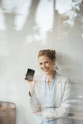 Happy businesswoman showing mobile phone in cafe seen through glass - JOSEF10816