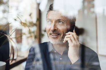 Smiling businessman talking on mobile phone seen through glass - JOSEF10832