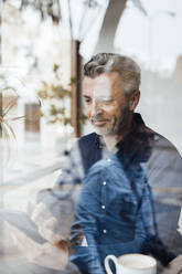 Smiling businessman using smart phone in cafe seen through glass - JOSEF10833