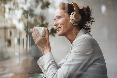 Lächelnde Geschäftsfrau mit Kaffeetasse, die über drahtlose Kopfhörer in einem Café Musik hört - JOSEF10876