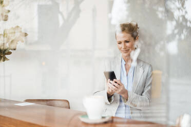 Smiling businesswoman looking at smart phone in cafe seen through glass - JOSEF10879