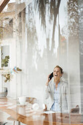 Cheerful businesswoman talking on mobile phone in cafe seen through glass - JOSEF10882