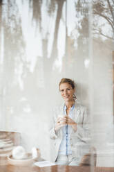 Lächelnde Geschäftsfrau mit Kaffeetasse im Café durch Glas gesehen - JOSEF10883