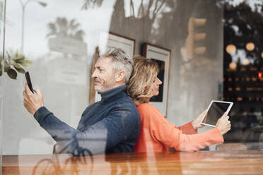 Smiling couple using wireless technologies sitting cafe seen through glass - JOSEF10903