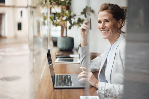 Happy businesswoman holding credit card sitting with laptop in cafe - JOSEF10915