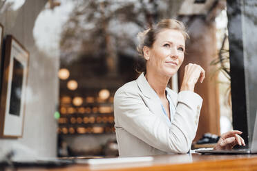 Smiling businesswoman sitting with laptop in cafe seen through glass - JOSEF10916