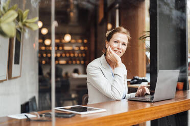 Smiling businesswoman sitting with head in hand at cafe seen through glass - JOSEF10921