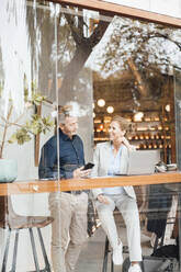 Lächelnde Geschäftsfrau mit Geschäftsmann, der ein Mobiltelefon in einem Café hält, gesehen durch ein Glasfenster - JOSEF10926