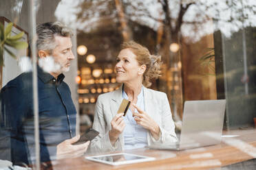 Businesswoman pointing at credit card discussing with businessman in cafe seen through glass - JOSEF10928