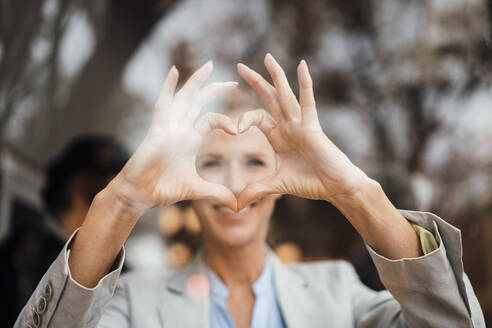 Smiling businesswoman gesturing heart shape in cafe seen through glass - JOSEF10937
