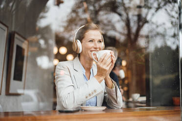 Lächelnde Geschäftsfrau mit Kaffeetasse, die über drahtlose Kopfhörer in einem Café Musik hört - JOSEF10938