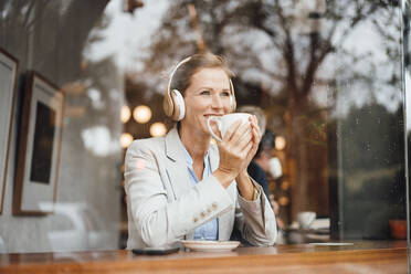 Smiling businesswoman holding coffee cup listening music through wireless headphones in cafe - JOSEF10938