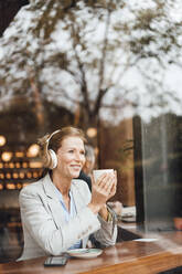 Smiling businesswoman holding coffee cup listening music in cafe seen through glass - JOSEF10939