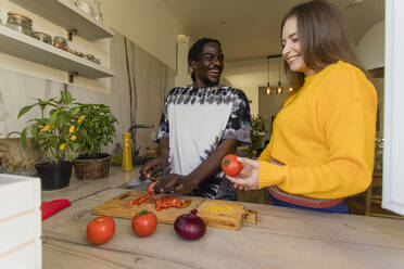 Smiling woman holding tomato standing by boyfriend cutting vegetables in kitchen - OSF00283