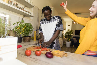 Glückliche Frau, die eine Tomate hält und neben ihrem Freund steht, der in der Küche Gemüse schneidet - OSF00284