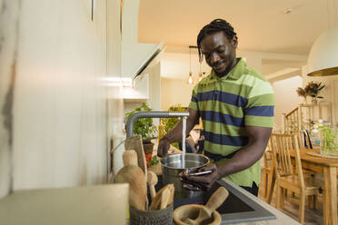 Lächelnder junger Mann, der einen Topf aus dem Wasserhahn in der Küche füllt - OSF00295
