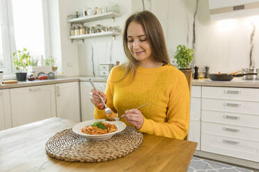 Smiling woman eating spaghetti sitting at dinning table at home - OSF00307