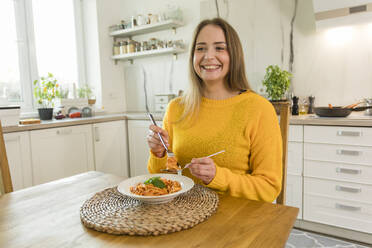 Happy woman eating spaghetti sitting at dinning table at home - OSF00308