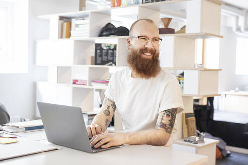 Lächelnder Mann mit Bart an einem Laptop am Tisch - FOLF11592