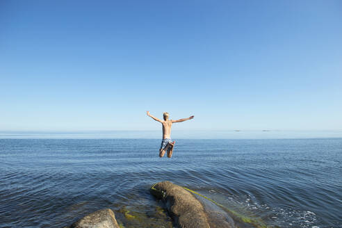 Young man jumping off rock by sea - FOLF11598