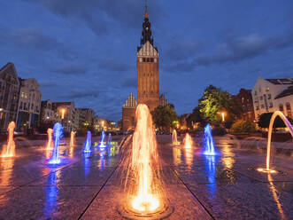 Poland, Warmian-Masurian Voivodeship, Elblag, Illuminated fountain at old town square with Saint Nicholas Cathedral in background - LAF02763