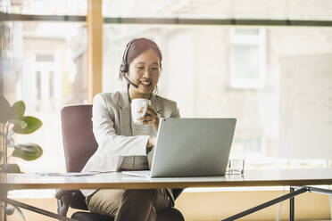 Happy businesswoman wearing headset working on laptop in office - UUF26726