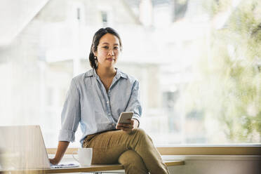 Smiling businesswoman with smart phone sitting on desk at work place - UUF26736