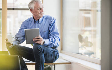 Senior businessman with tablet PC looking through window in office - UUF26748