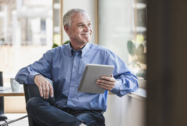 Happy businessman with tablet PC sitting by window - UUF26749