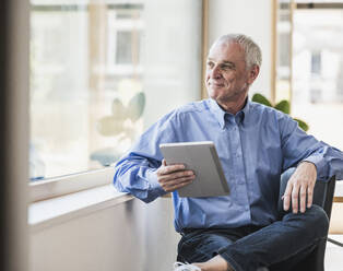 Smiling senior businessman with tablet PC sitting by window - UUF26750