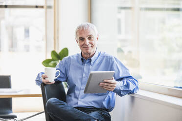 Smiling businessman with coffee cup and tablet PC at work place - UUF26751