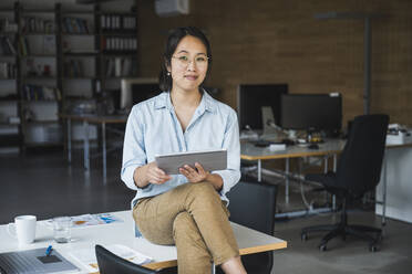 Smiling businesswoman wearing eyeglasses sitting on desk at work place - UUF26783