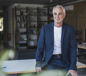 Lächelnder älterer Geschäftsmann sitzt auf einem Schreibtisch im Büro - UUF26790