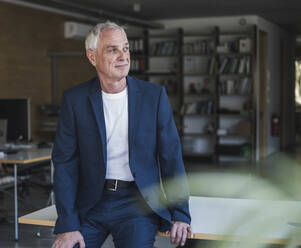 Smiling businessman sitting on desk in office - UUF26792