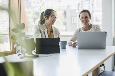 Happy mature businesswoman with colleague discussing over laptop in office - UUF26815