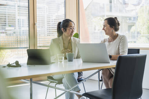 Smiling mature businesswoman with colleague discussing over laptop in office - UUF26816