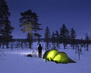 Young woman standing by tent in snowy forest at night - FOLF11779