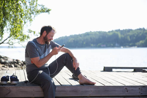 Mann benutzt sein Smartphone, während er auf dem Bootssteg sitzt - FOLF11895