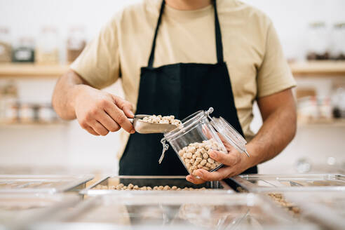 Anonymous shopkeeper pouring raw chickpeas inside glass jar in local store - ADSF35613