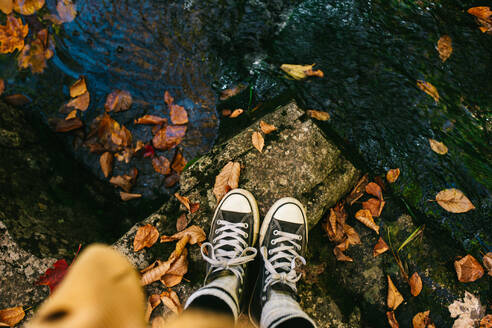 From above unrecognizable person foot standing on rock surface near brook full of maple yellow leaf - ADSF35621