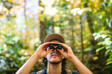 Junger männlicher Rucksacktourist, der im Wald steht und mit einem Fernglas den Waldgipfel beobachtet, während einer Wanderung im Sommer - ADSF35623