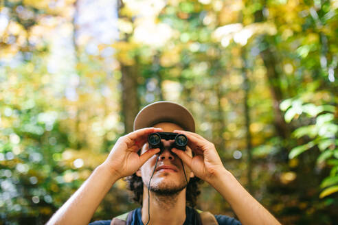 Young male backpacker standing on forest observing wood top in binoculars during trekking in summer - ADSF35623